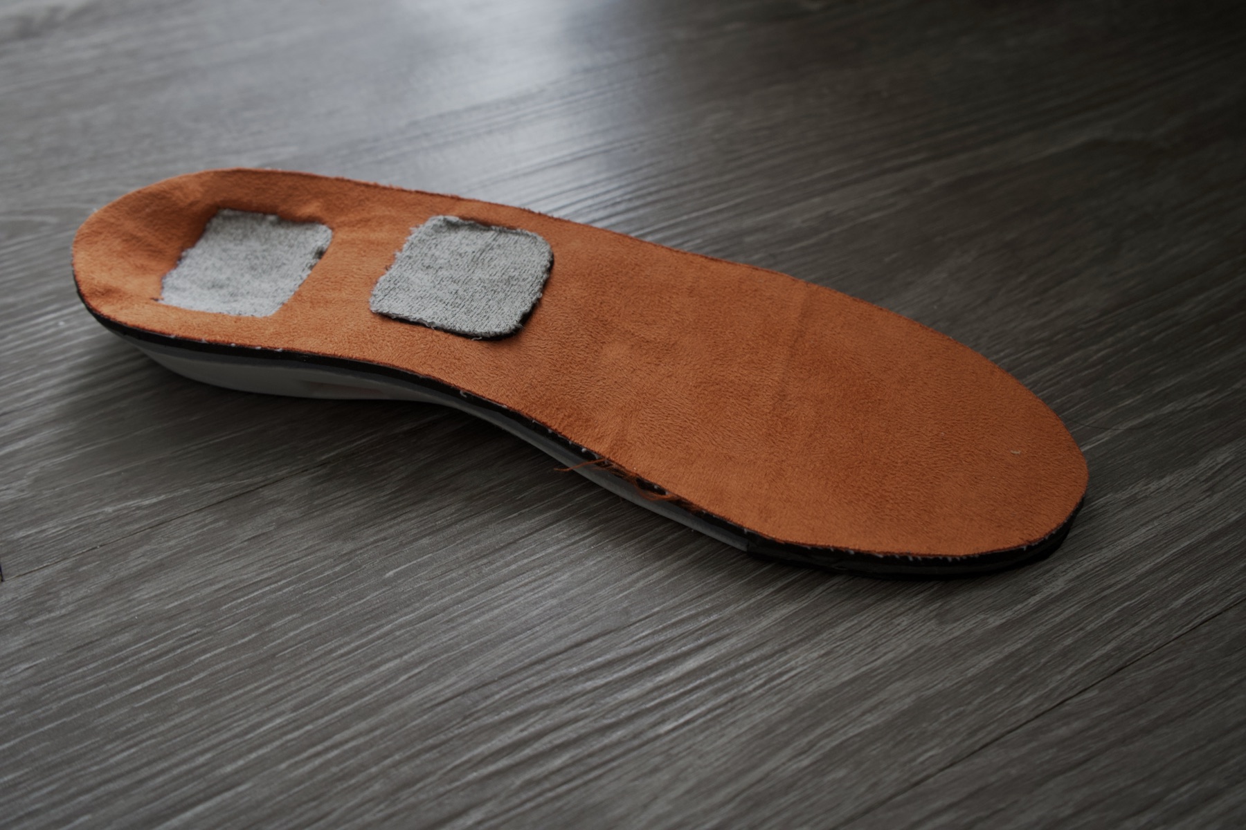 The NeuraSole V1 prototype photographed on a dark wood floor, showing the tan top cover and two conductive fabric patches at the heel and forefoot